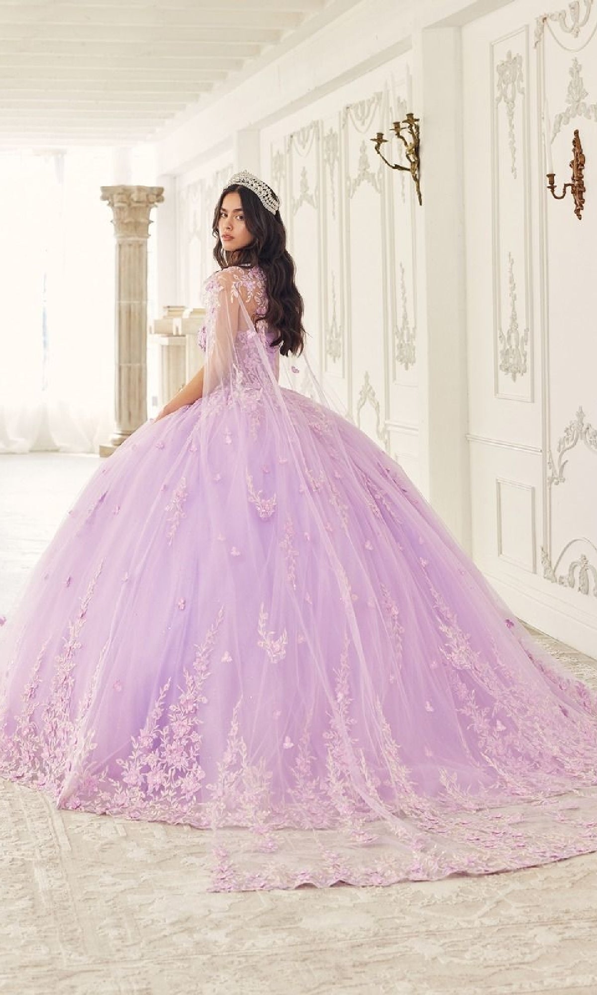 A woman in the Quinceanera Dress 15723 by Ladivine, featuring lavender floral lace applique and a corset bodice, stands in an ornate white room with a tiara, facing partially away from the camera.